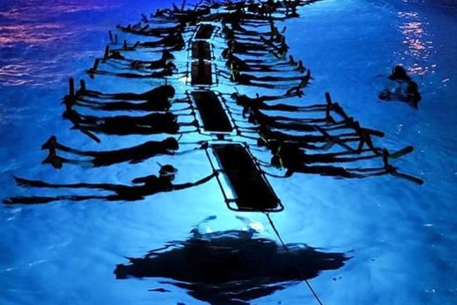 People lying on floating mats in a pool, illuminated by blue lighting.