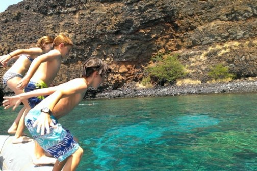 Three people diving into clear blue water from a boat near rocky cliffs.