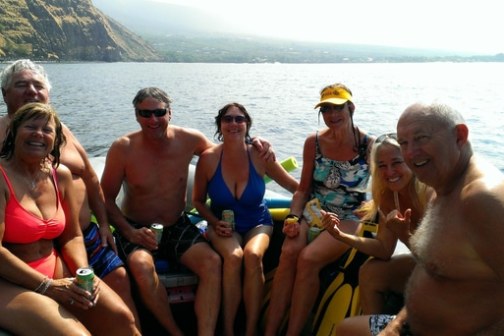 Group of seven people in swimsuits smiling on a boat on a sunny day.