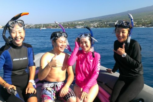 Four people with snorkels smiling on a boat, near the coast, with a scenic background.