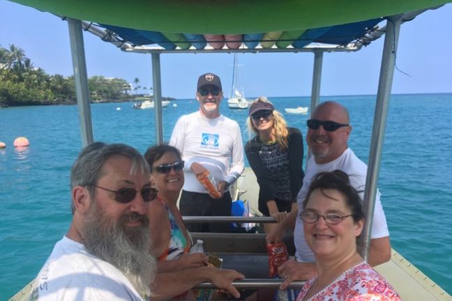 Six people smiling on a boat with the ocean and sailboats in the background.