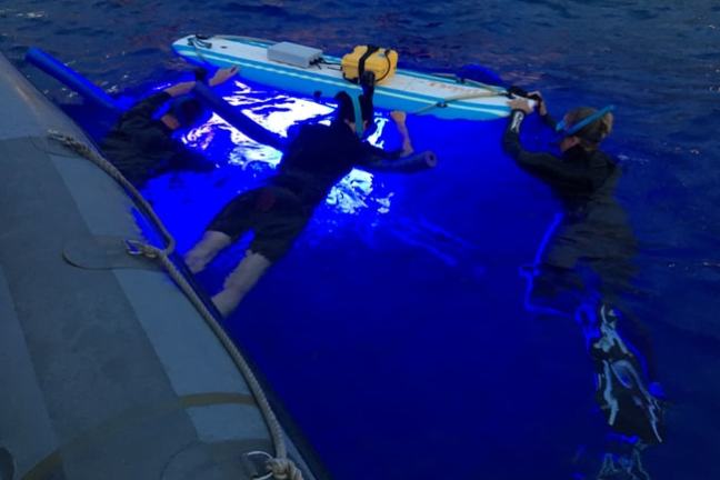Three people in wetsuits float near a boat, illuminated by blue light, holding a surfboard with gear.
