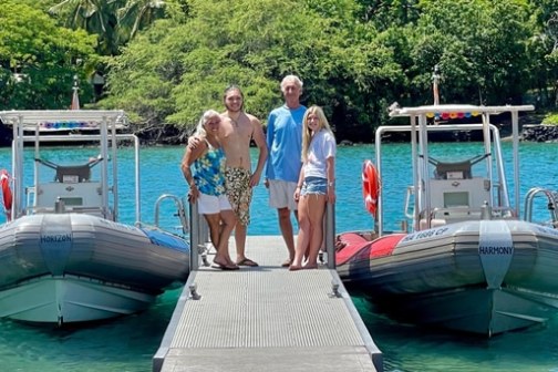 Four people stand on a dock between two boats with lush greenery in the background.
