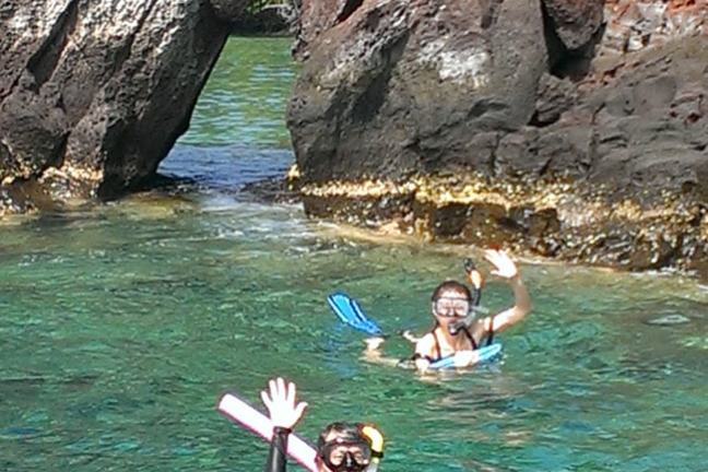 Two snorkelers in clear water near rocky cliffs, waving at the camera.