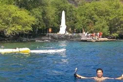 Man swimming near kayak with monument and trees in the background.