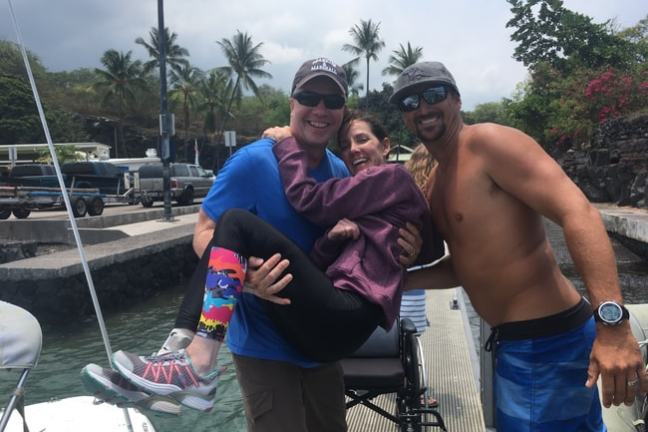 Three people smiling, one man carrying a woman near a dock with palm trees in the background.