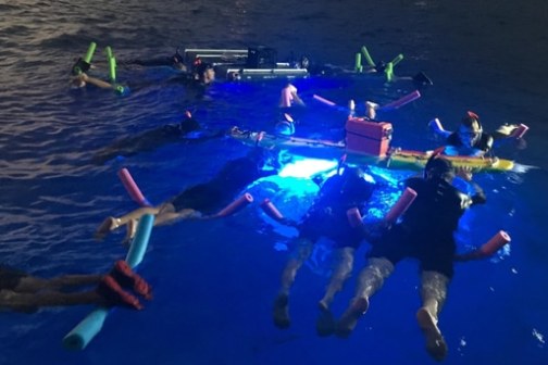 People floating in water at night with pool noodles and bright blue lights