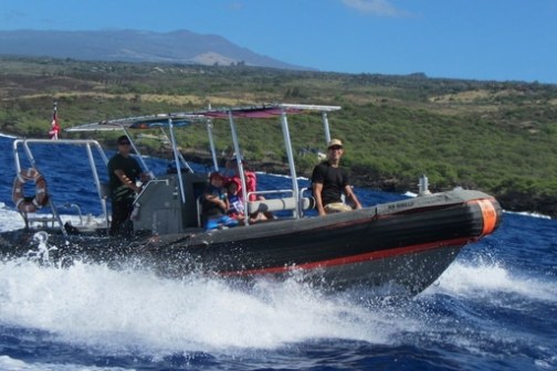 People on a small motorboat speeding through blue water near a green coastline.