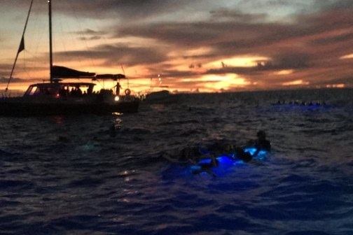 Sailboat at sunset with swimmers and glowing lights in the ocean.
