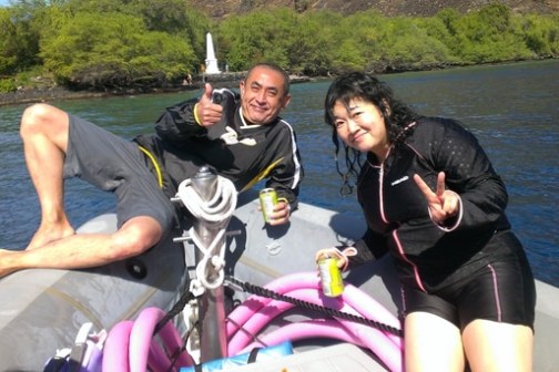 Two people on a boat with drinks, making peace signs, near lush shoreline.