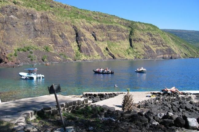 Boats on a calm bay near rocky cliffs, with people sunbathing on a rocky shore.