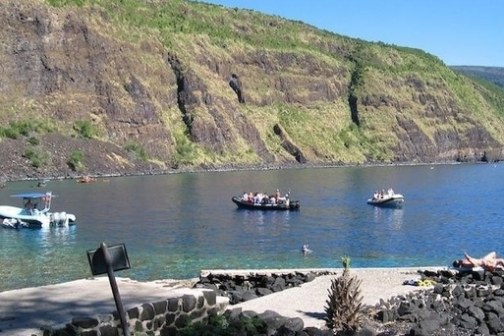 Three boats on a blue lake beneath green cliffs with sunbathers on rocky shore.