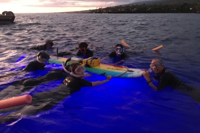 People snorkeling at sunset, holding a surfboard with lights in the water.