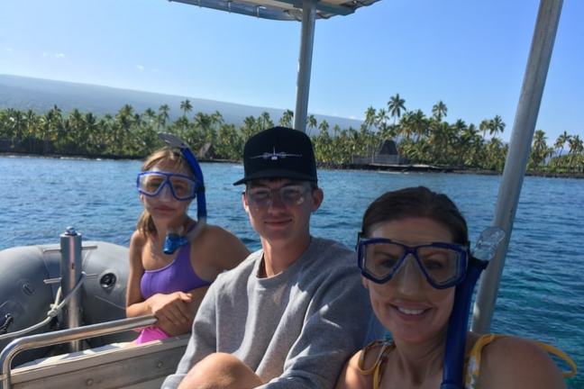 Three people in snorkeling gear sit on a boat, tropical island in background.